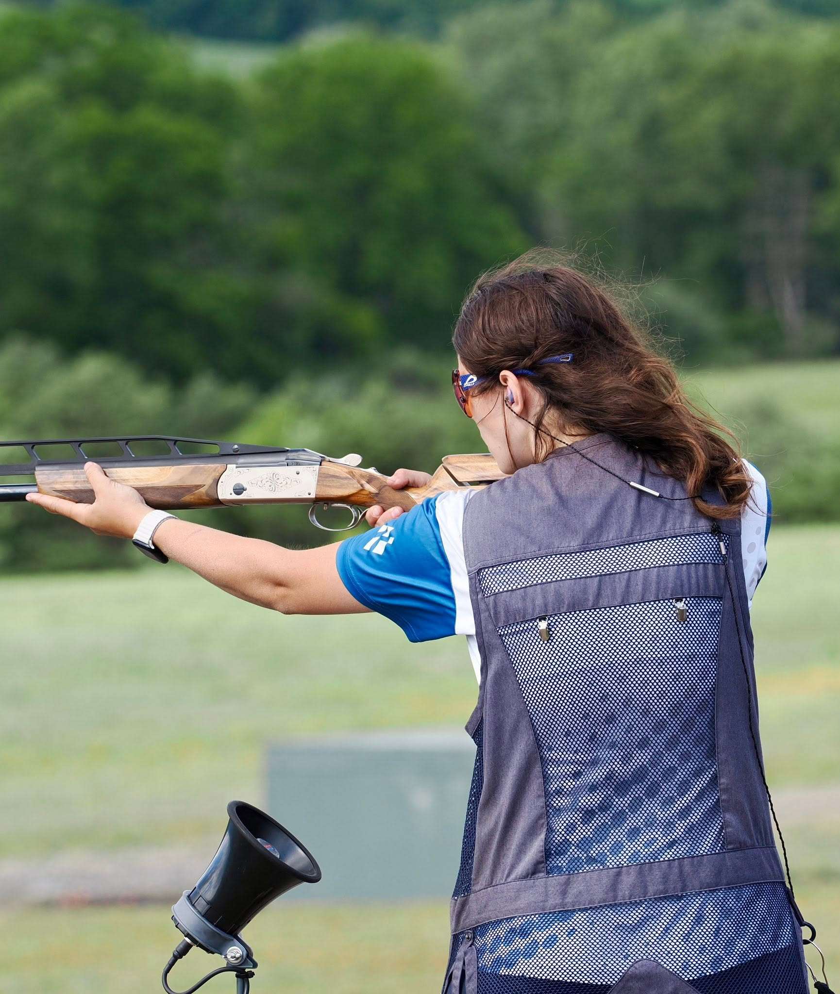 Clare Schaffer in a shooting vest and protective glasses shoulders an engraved over-under shotgun on an outdoor trap range, viewed from behind with a green tree line in the background