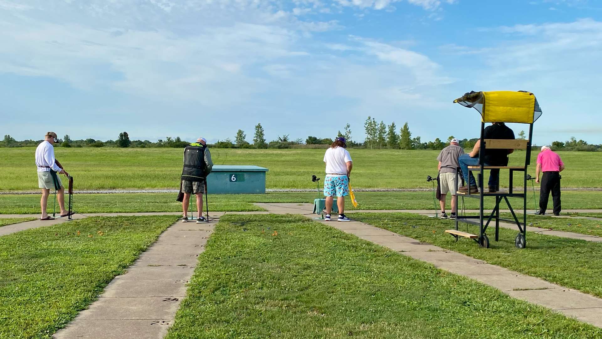 Five trapshooters stand at their stations on trap field 6 at the Grand American, guns pointed downrange toward the green trap house, with a referee seated in a yellow-canopied chair at right