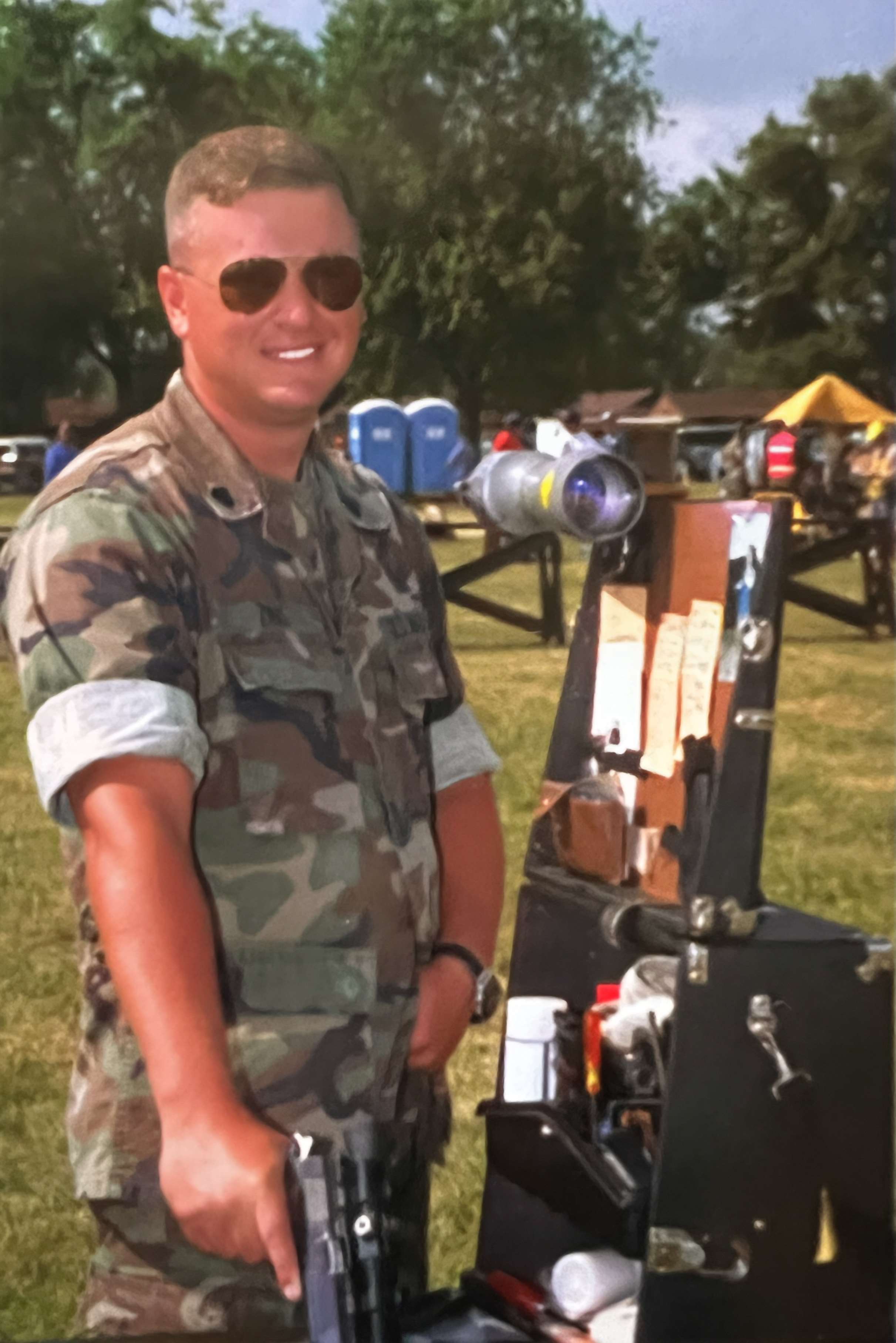 Brian Zins, in woodland camouflage utilities and aviator sunglasses, stands beside a spotting scope and pistol box on the firing line