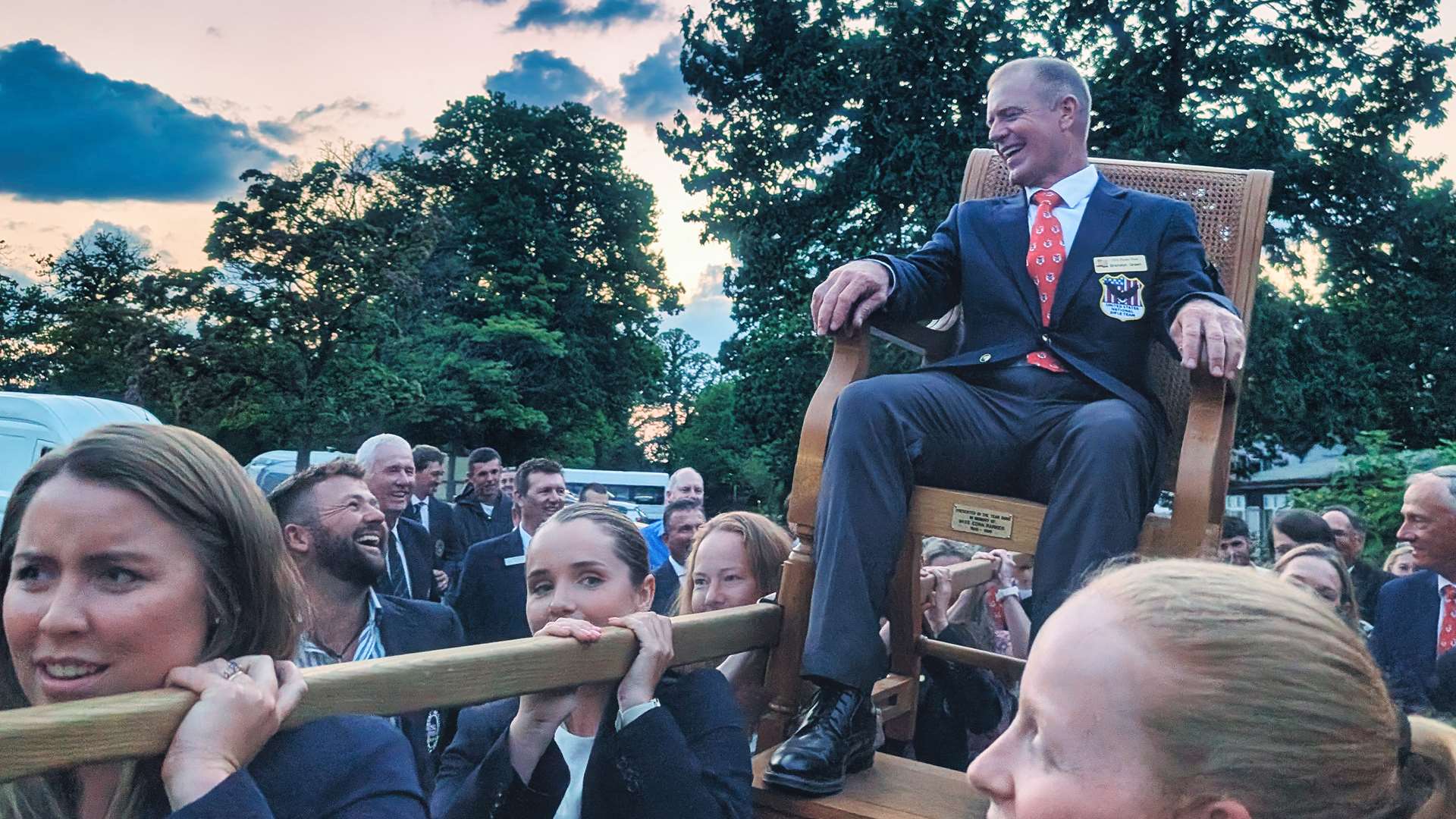 Brandon Green sits in a wooden ceremonial chair held aloft on poles by teammates in dark blazers, smiling against a dusk sky at Bisley Camp in England
