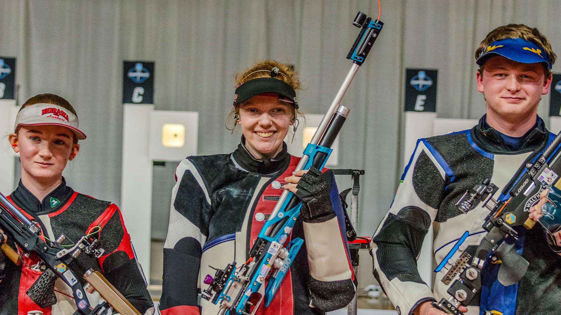 Three college rifle shooters in shooting jackets hold air rifles and smile after the air rifle final at the 2026 NCAA Rifle Championship