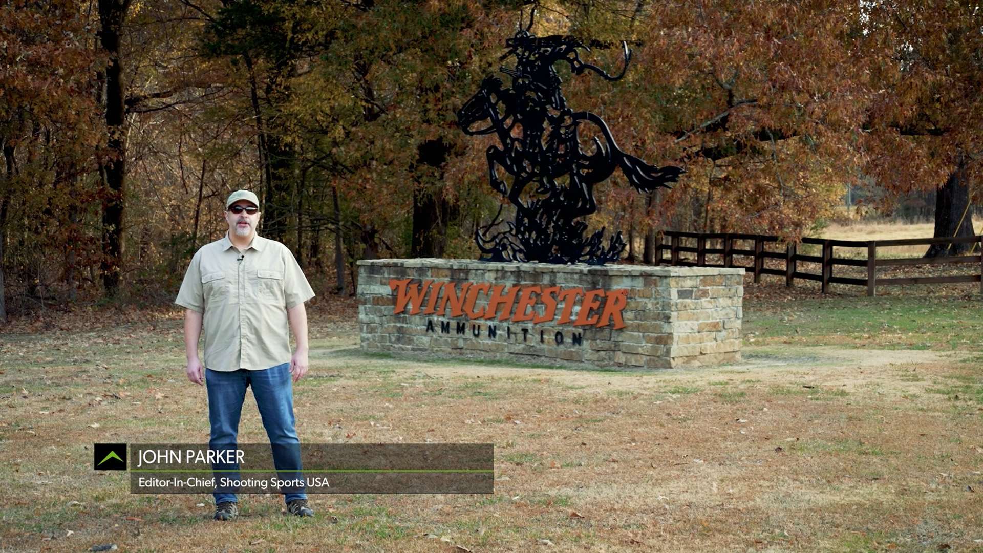 John Parker, editor-in-chief of Shooting Sports USA, standing in front of the Winchester Ammunition entrance sign featuring a black metal horse-and-rider sculpture surrounded by autumn trees