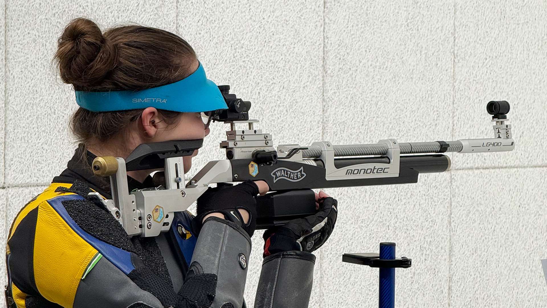 Close-up of a female rifle athlete in a blue visor and blue-and-gold shooting jacket aiming a Walther air rifle from the standing position