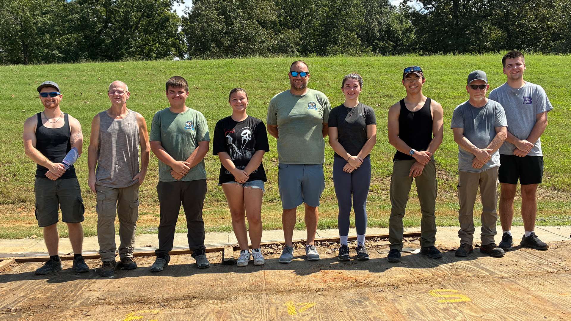 Nine people, most college-aged, standing shoulder to shoulder and smiling on a concrete shooting pad marked with yellow stenciled lane numbers, with a green grass berm rising behind them