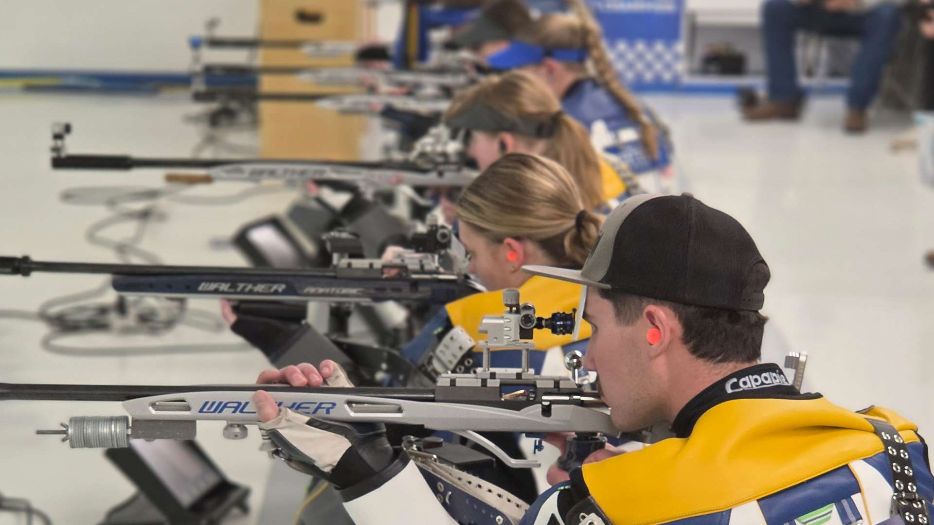 A row of college rifle shooters lies in the prone position aiming smallbore rifles, with electronic scoring monitors on the floor beside them