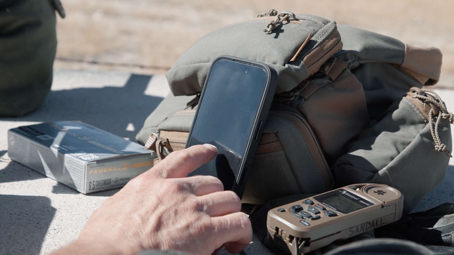 Hand tapping a smartphone propped on a green range bag, next to a box of Federal 6.5 Creedmoor +Peak ammunition and a Kestrel meter