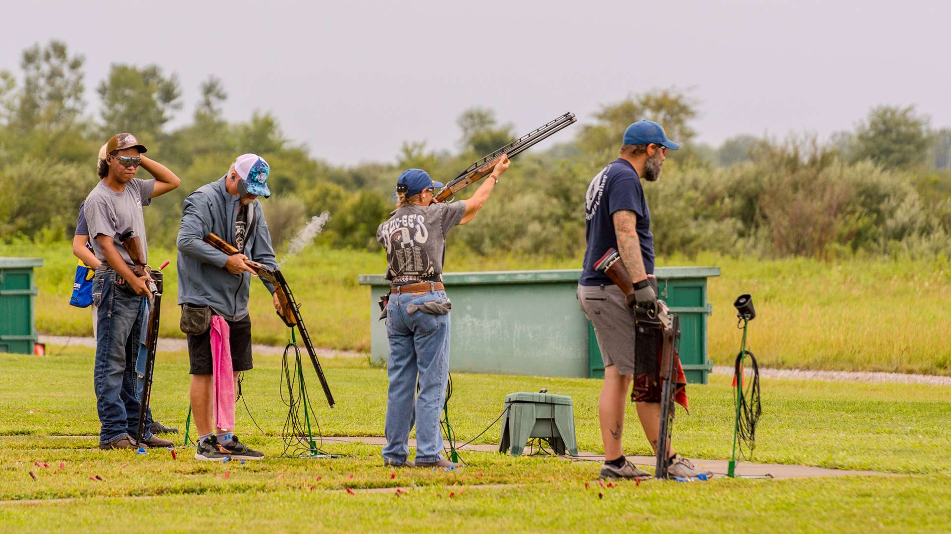 Four trapshooters on a station during the Grand American; one woman fires while smoke drifts from a nearby shooter's open action