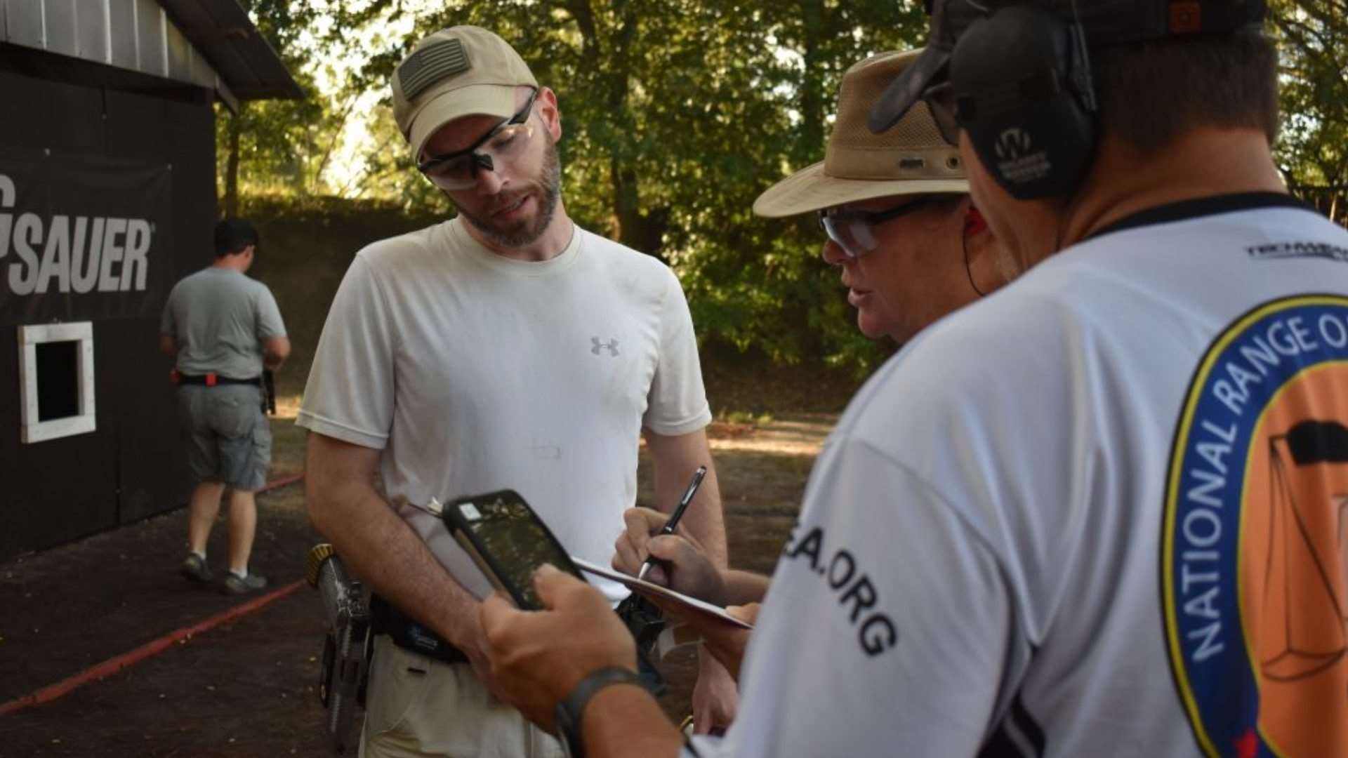 A competitor in a tan cap and gray shirt stands talking with two Range Officers, one wearing a National Range Officers Institute jersey, while one of the ROs writes on a scoresheet held on a clipboard