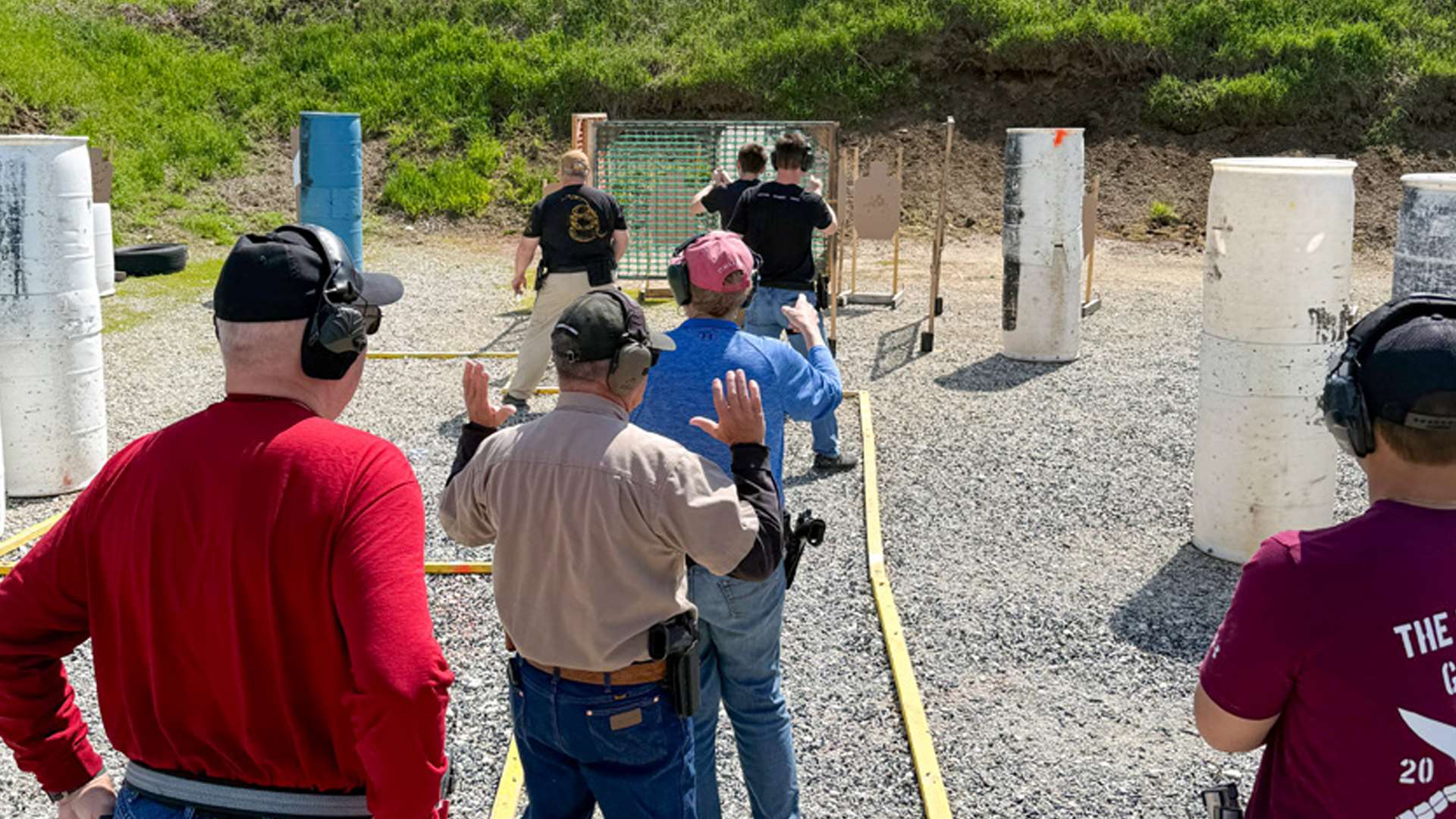 USPSA competitors with raised hands rehearse a stage plan during a walkthrough on a gravel bay lined with white barrels and cardboard targets