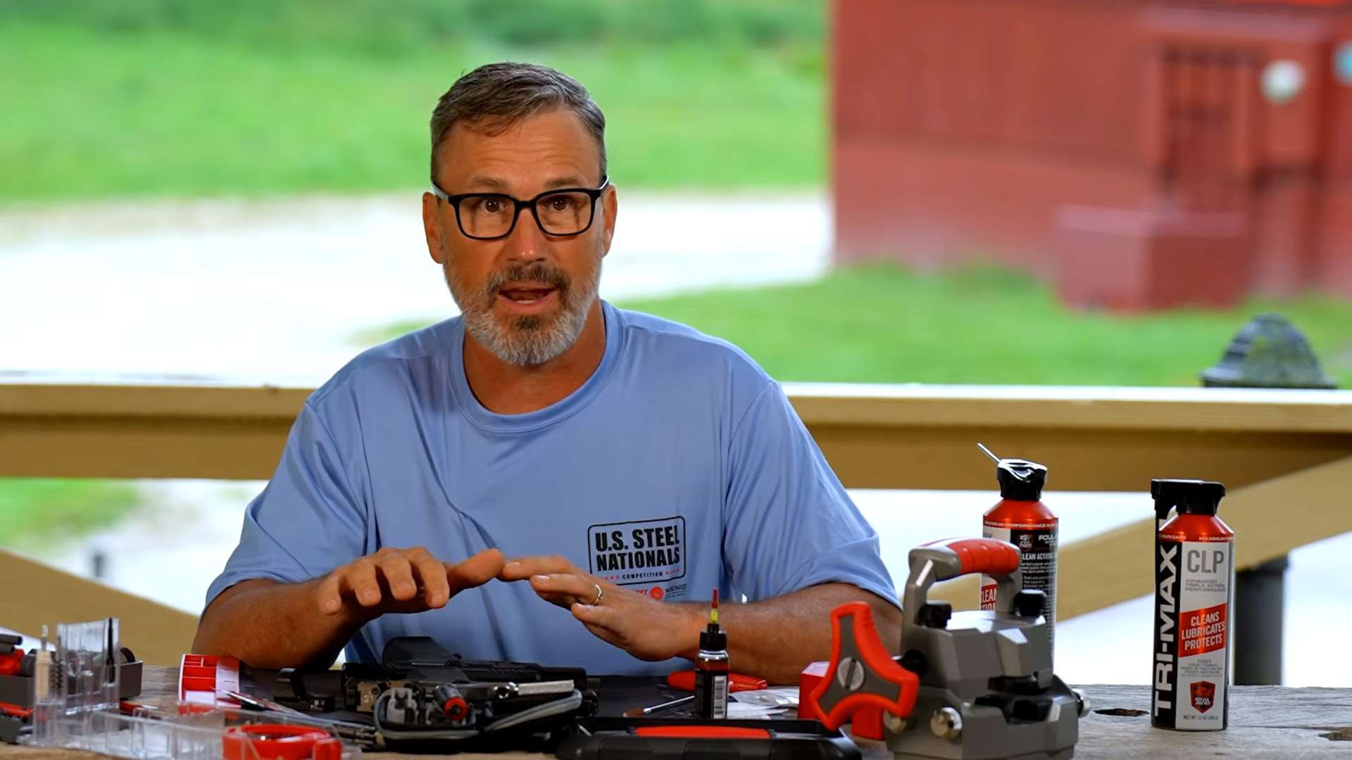 Doug Koenig seated at an outdoor range bench with a disassembled pistol, Real Avid cleaning tools, a bench vise and a can of Tri-Max CLP in front of him