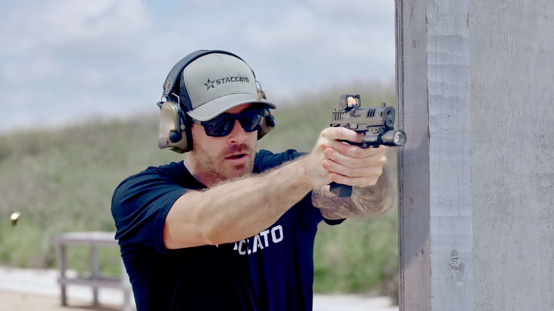 Shooter wearing Staccato hat and ear protection firing the HD P4X pistol with red-dot optic and light at an outdoor range beside a barricade