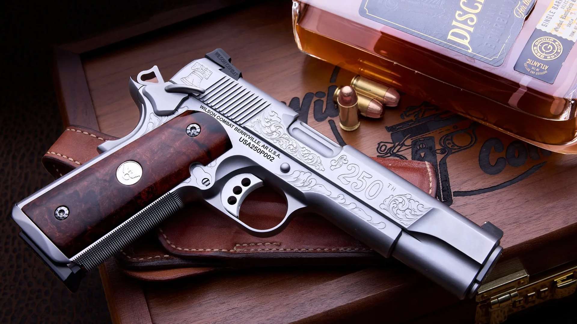 Stainless steel Wilson Combat America 250th 1911 pistol with hand-engraved scrollwork, 250th slide marking and Desert Ironwood grips, resting on a leather holster beside cartridges