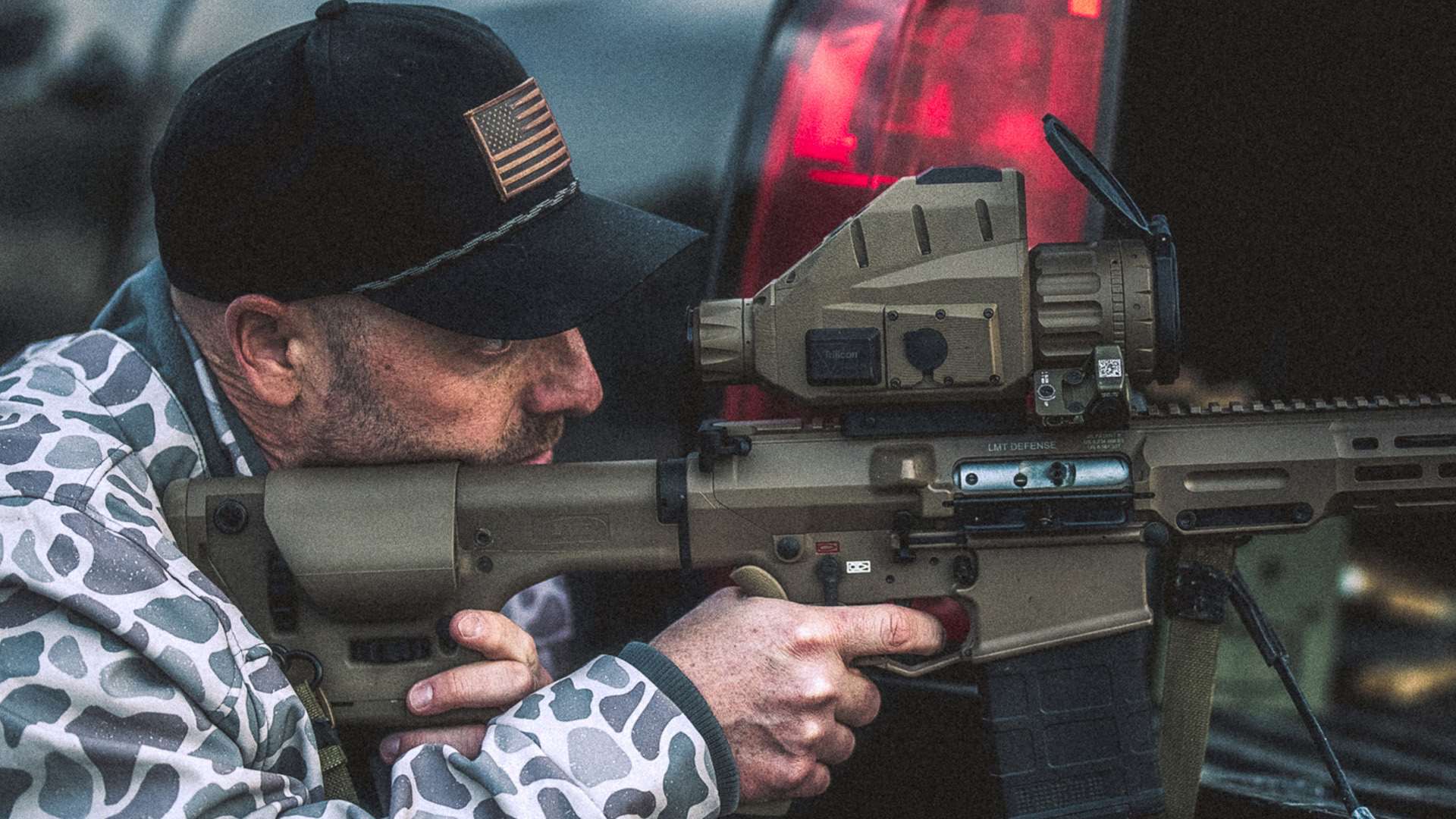 Man in a black cap with a flag patch and gray camo jacket aims a tan LMT Defense rifle fitted with a Trijicon REAP-IR-50LRF thermal scope
