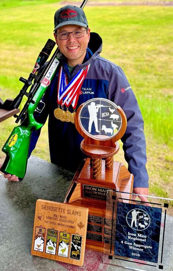 Close-up of a smiling Erich Mietenkorte in a Team Lapua jacket and gold medals, a green-stocked rifle with a Lapua-branded scope over his shoulder and the HCSA Iron Man trophy in front of him