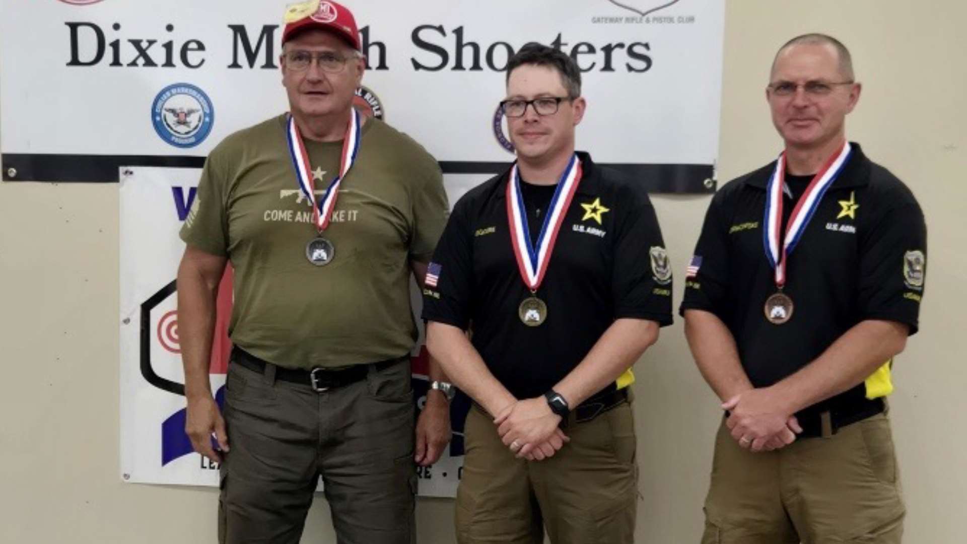 Three medalists stand in front of the Dixie Match Shooters banner after the Distinguished Revolver Match, with Randy Erickson at left in a Come and Take It t-shirt alongside Jason Gregoire and Greg Markowski in U.S. Army polos
