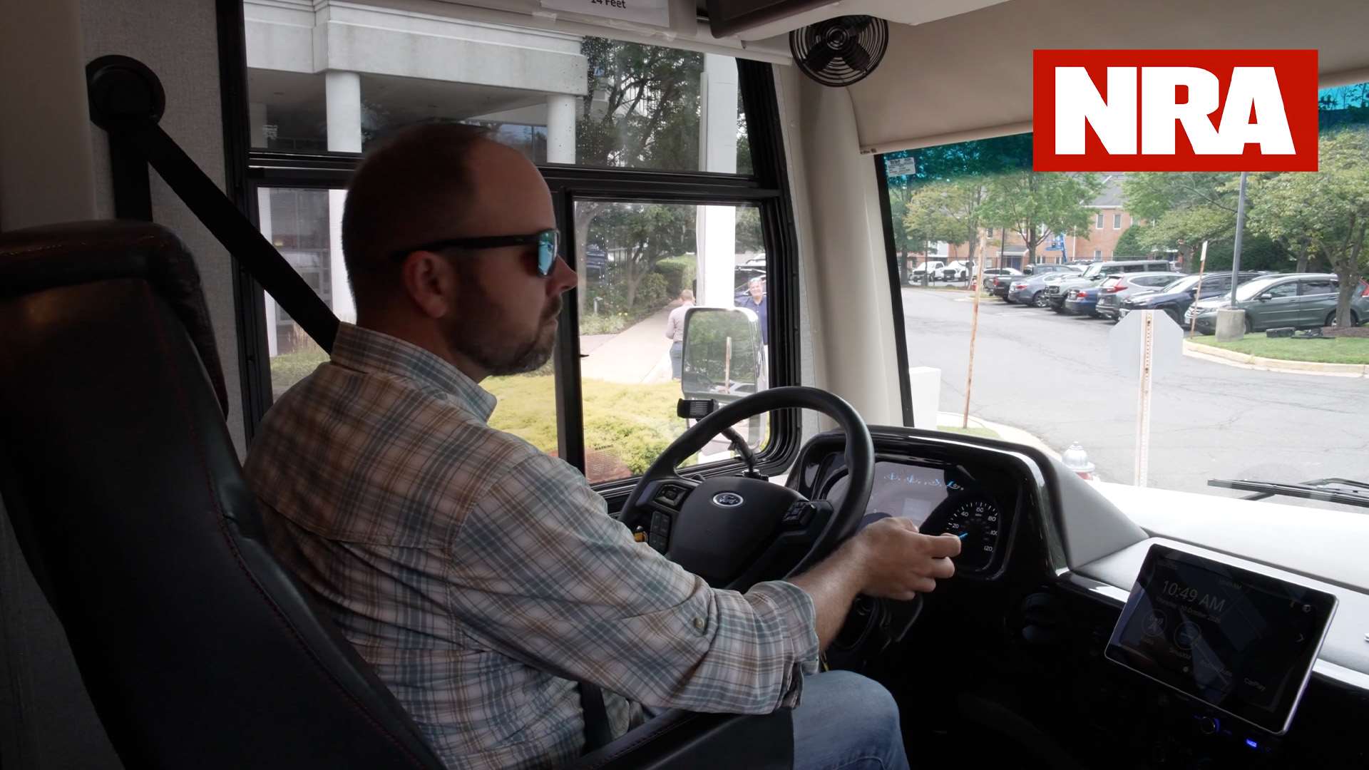 Man in sunglasses and a plaid shirt driving the NRA Gun Bus from the coach's front cab, headquarters grounds visible through the windshield