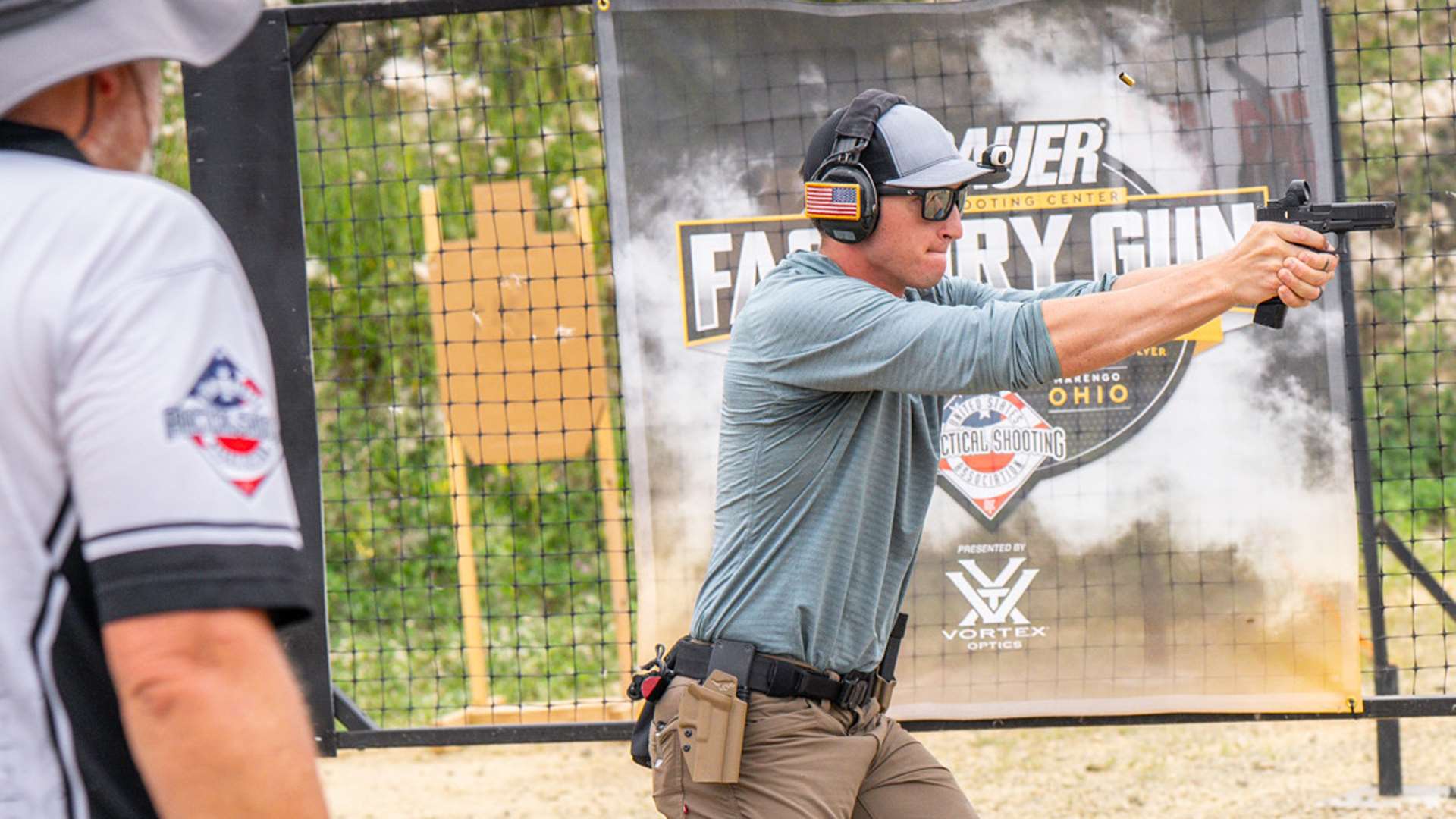 USPSA competitor in a gray shirt fires a pistol mid-stage while a Range Officer observes from the left, with a Factory Gun Nationals banner and netting in the background