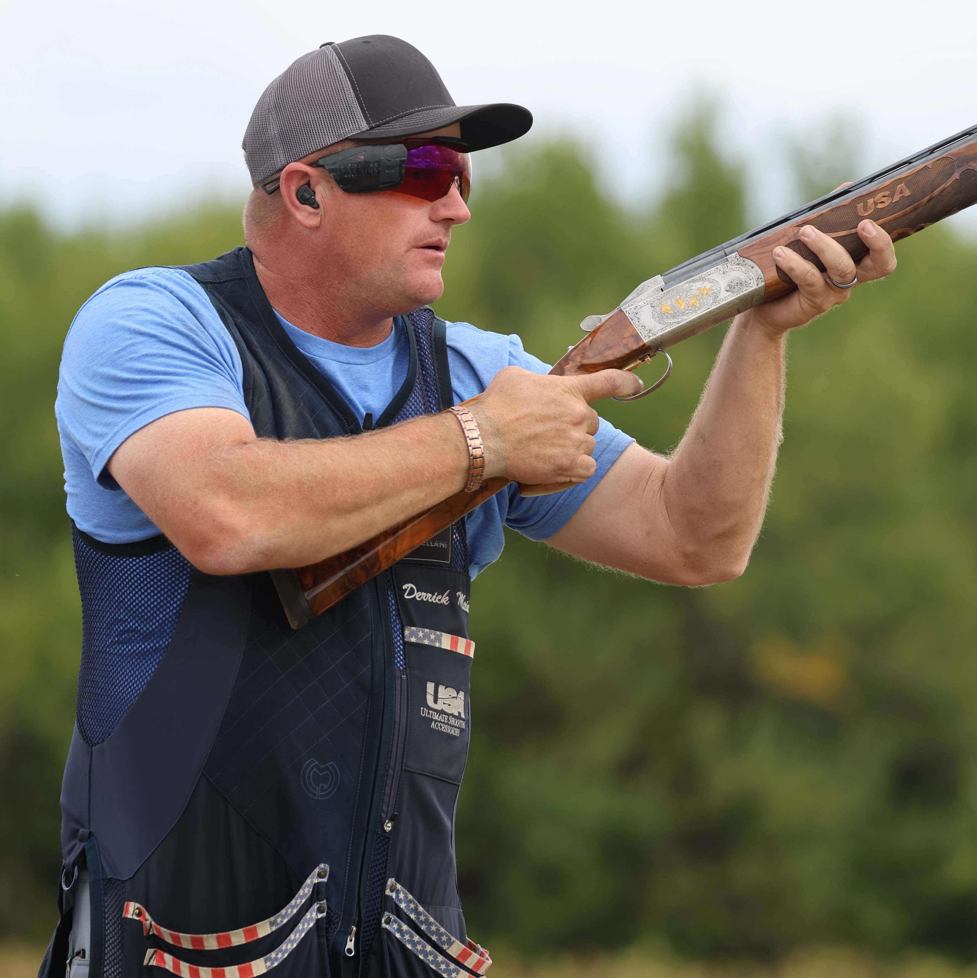 Derrick Mein, wearing a navy shooting vest embroidered with his name, holds an over-under shotgun with a carved “USA” fore-end while waiting to call for a target