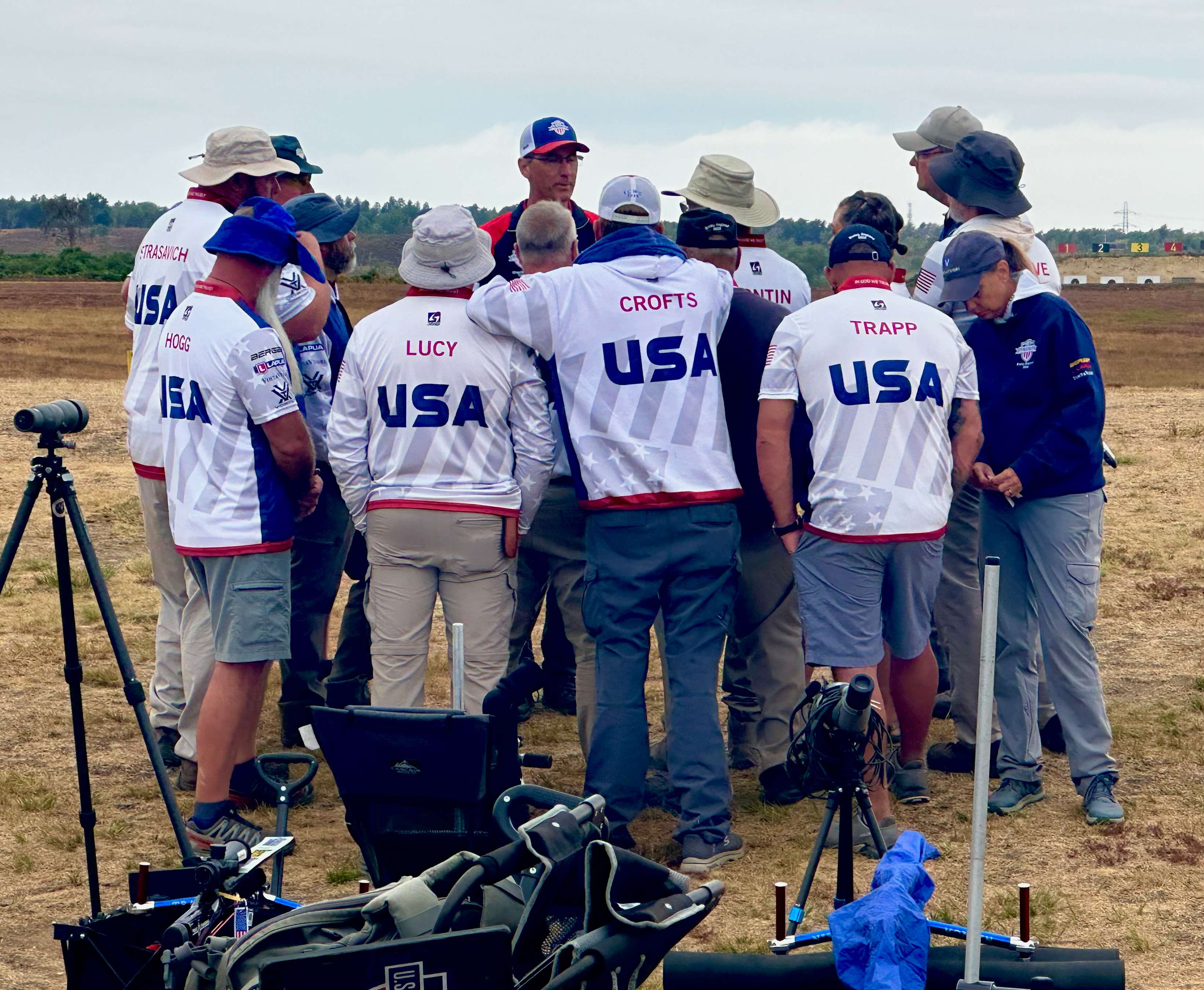 A U.S. F-T/R team huddle on the range at Bisley, shooters in white USA jerseys gathered around their captain with spotting scopes and equipment carts in the foreground and target butts in the distance