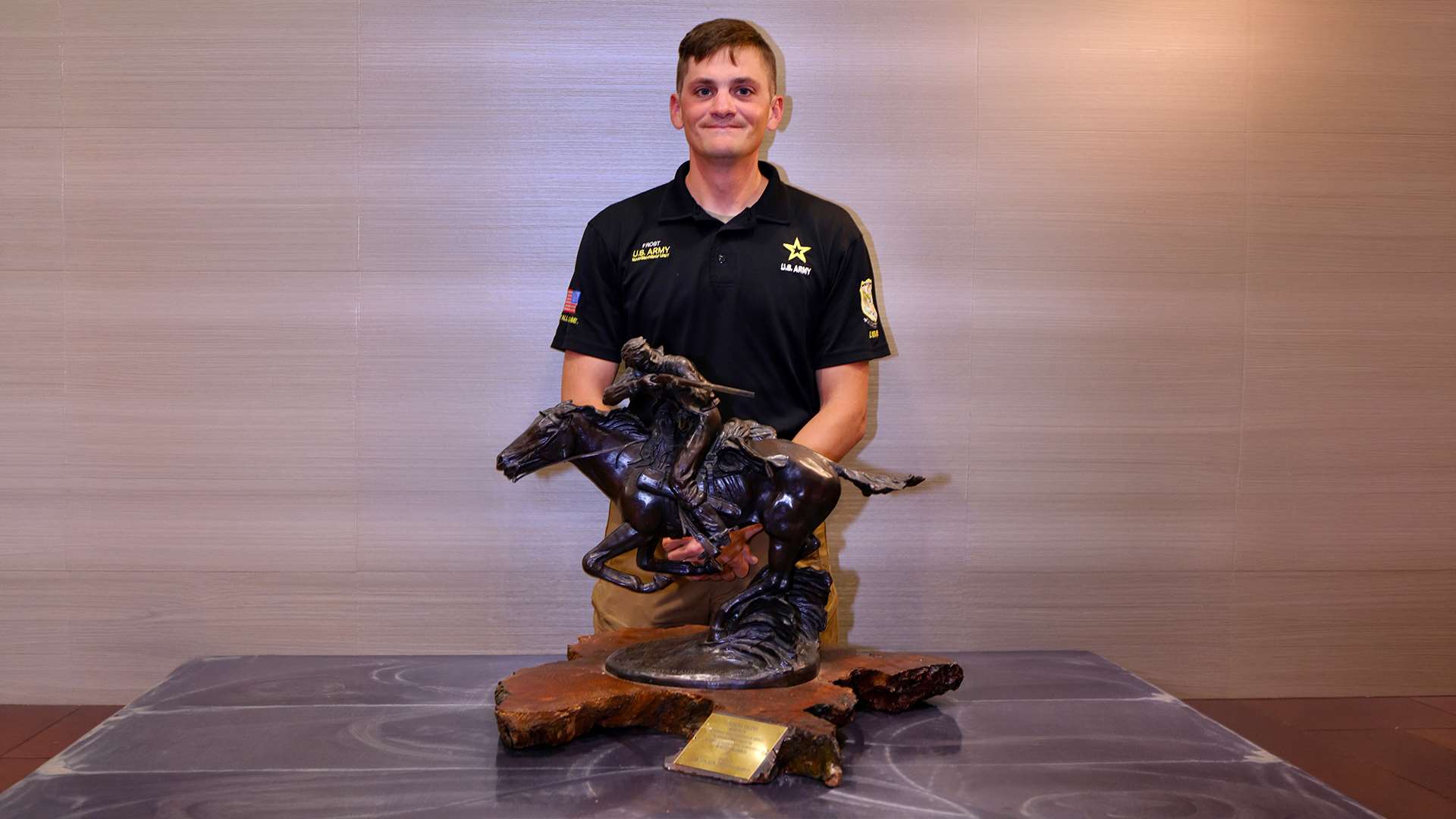 Logan Frost in a U.S. AMU polo shirt poses behind the Tompkins Trophy, a bronze sculpture of a mounted horseman on a natural wood base