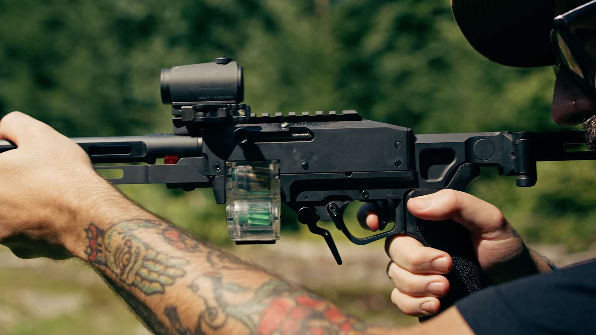 Close-up of the left side of a Tandemkross ADX Race Rifle receiver showing the left-side charging handle, integral Picatinny rail, Aimpoint Micro T-1 red dot and transparent magazine, with a shooter's hands on the fore-end and grip