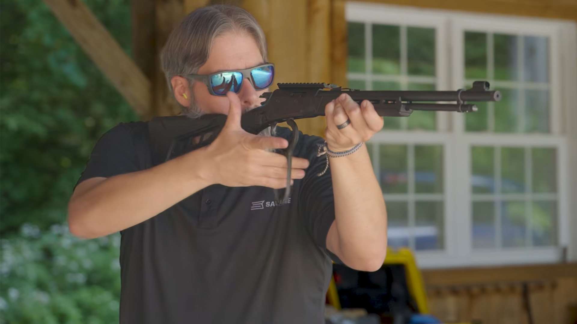 Man in sunglasses and a black Savage polo shirt shouldering a black lever-action rifle and cycling the lever at an outdoor range
