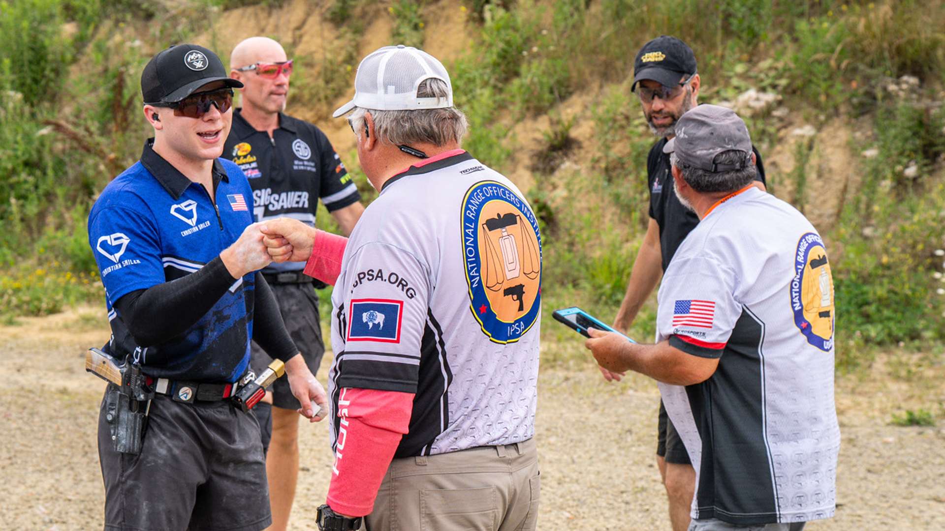 A Range Officer in a National Range Officers Institute jersey fist-bumps a competitor in a blue jersey while other match officials review a tablet on a gravel range