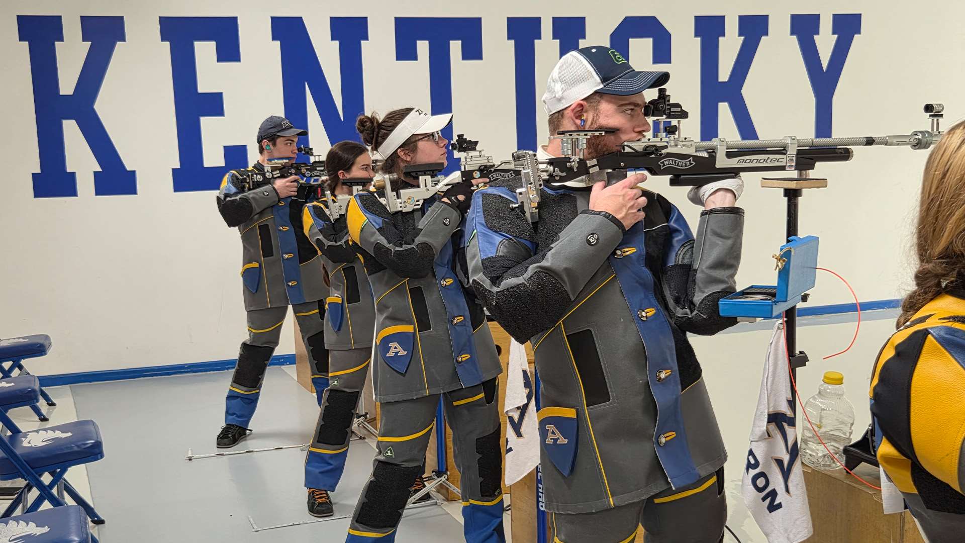 Four college rifle shooters in gray, blue and gold shooting jackets aim air rifles from the standing position in front of a white wall painted with large blue letters spelling KENTUCKY