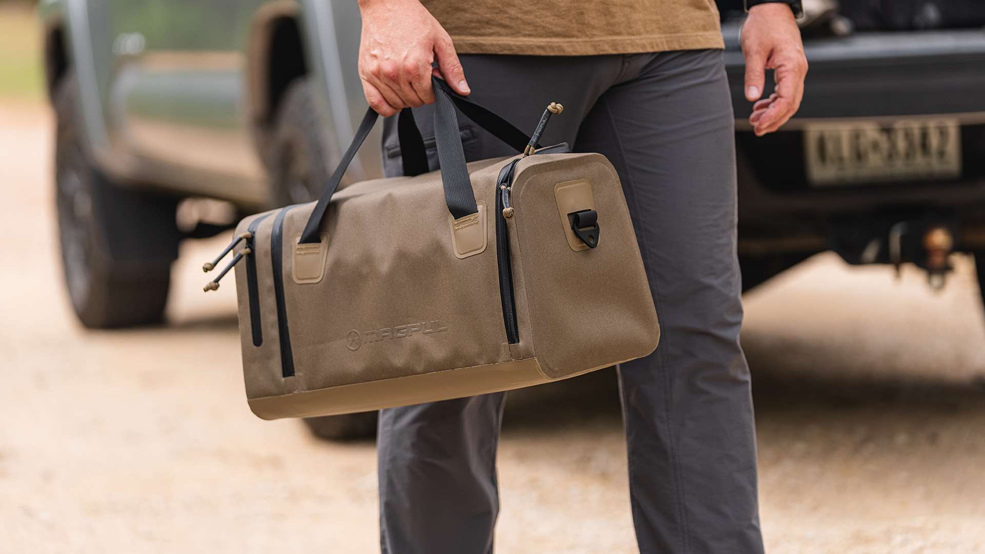 Bag holder carrying the flat dark earth Magpul DAKA Range Bag, Small, by its webbing handles beside a pickup truck on a gravel road