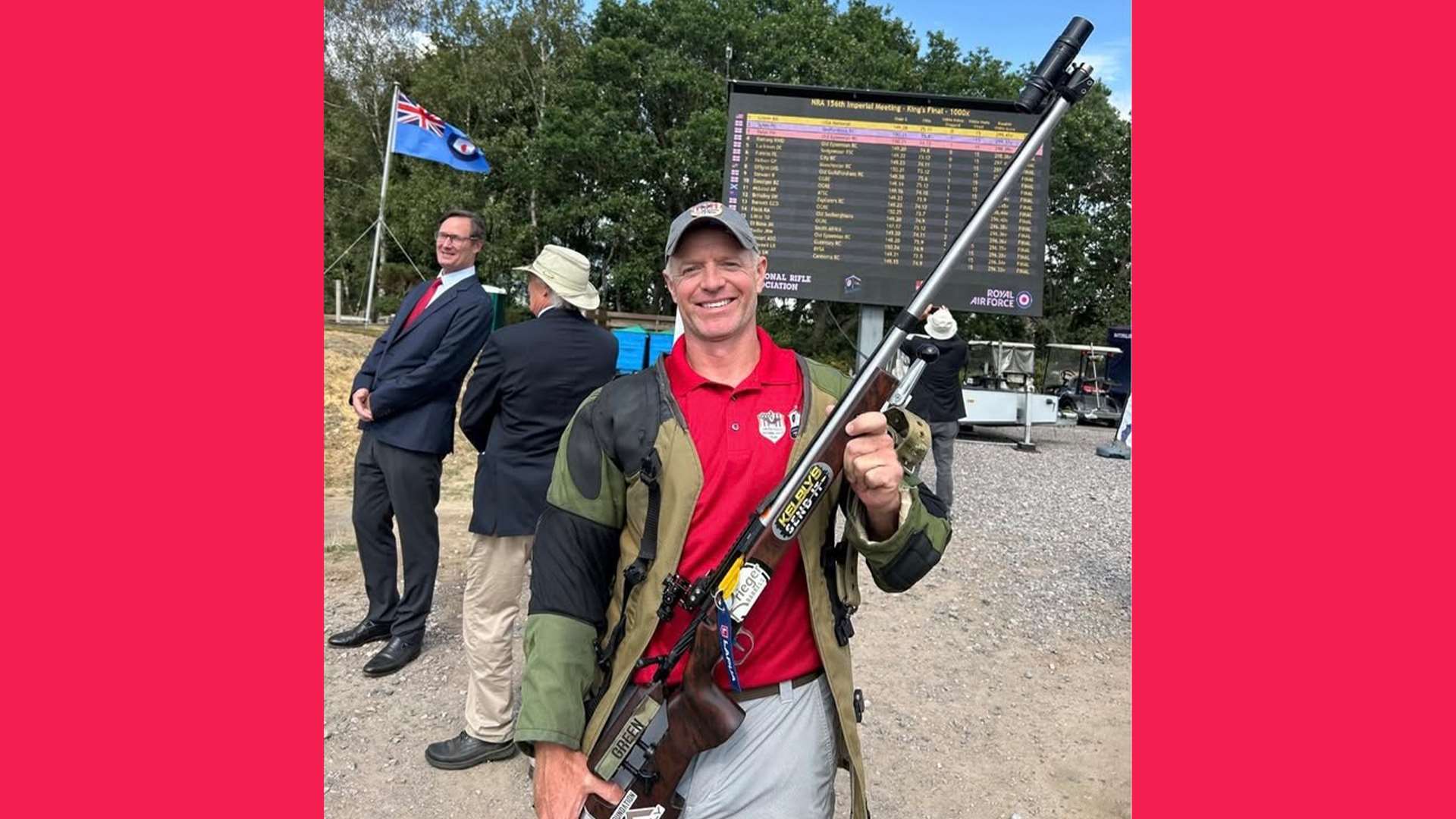 Brandon Green in a red shirt and shooting jacket holds his iron-sighted target rifle in front of an electronic scoreboard showing the King's Final results at Bisley