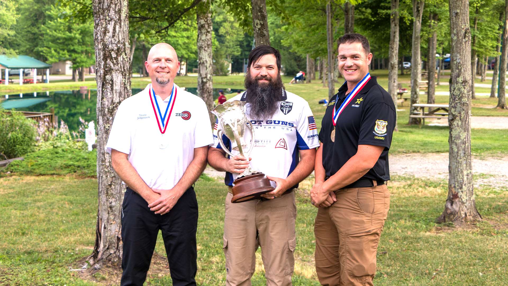 Travis Jorgenson, Jon Shue holding the Harrison Trophy and Ryan Franks pose outdoors among trees at the Cardinal Shooting Center after the 2025 NRA National Precision Pistol Championship awards ceremony