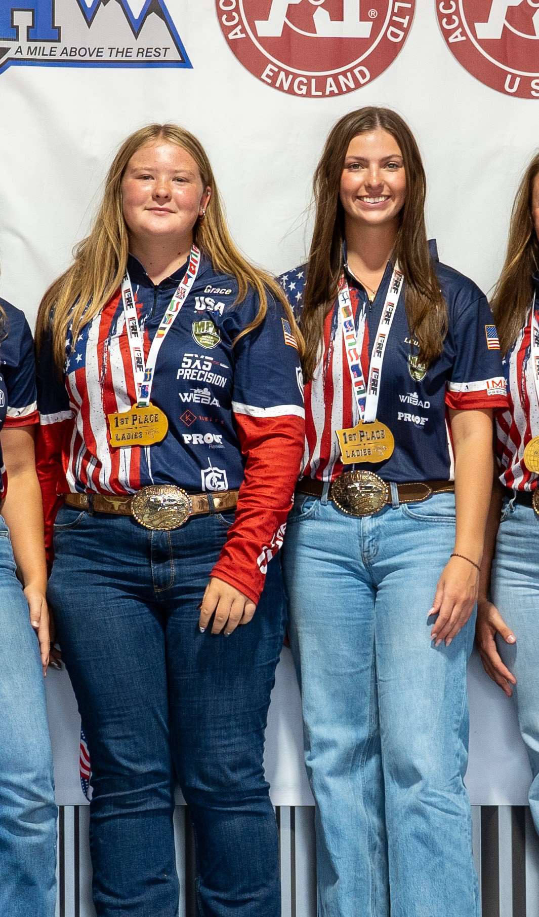 Two smiling female precision rifle shooters in red, white and blue Team USA jerseys stand side by side wearing gold first-place Ladies medals and trophy belt buckles