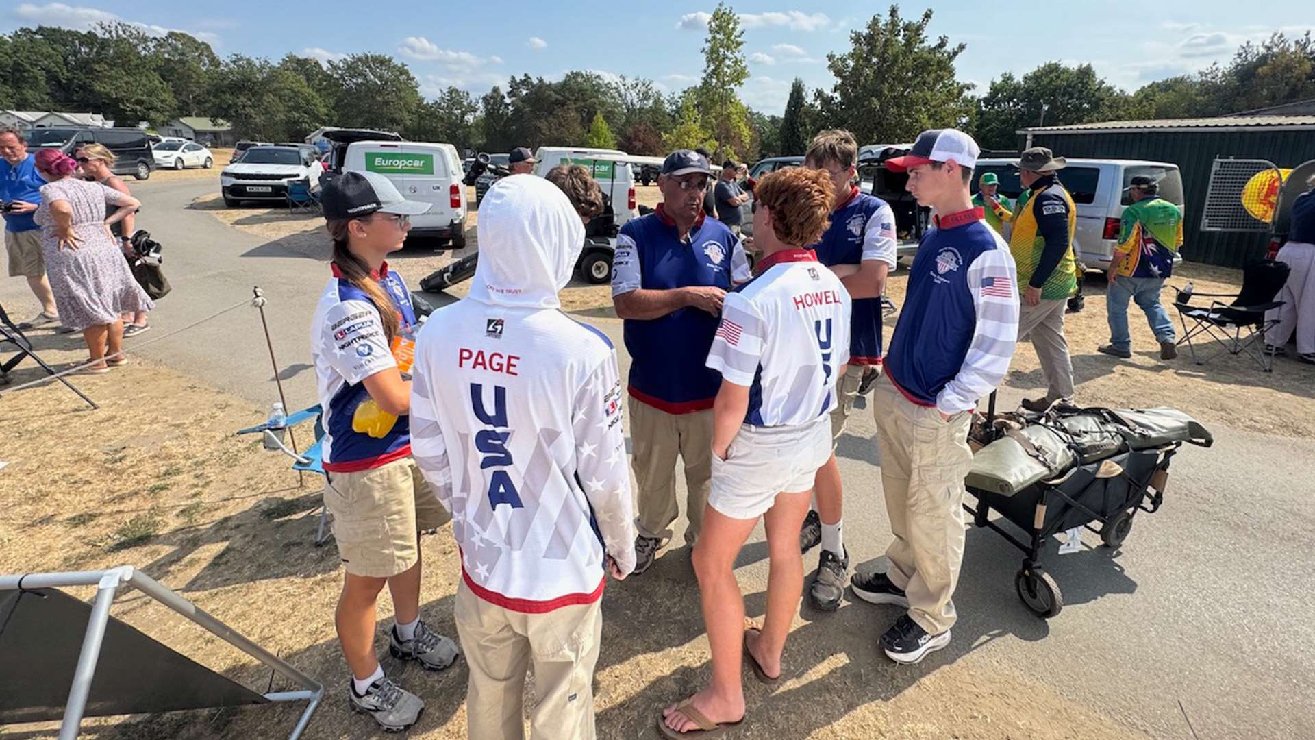 A group of young American F-Class competitors in USA team jerseys standing in a loose circle with an adult team official in a parking area behind the range at Bisley, two of the jerseys marked Page and Howell