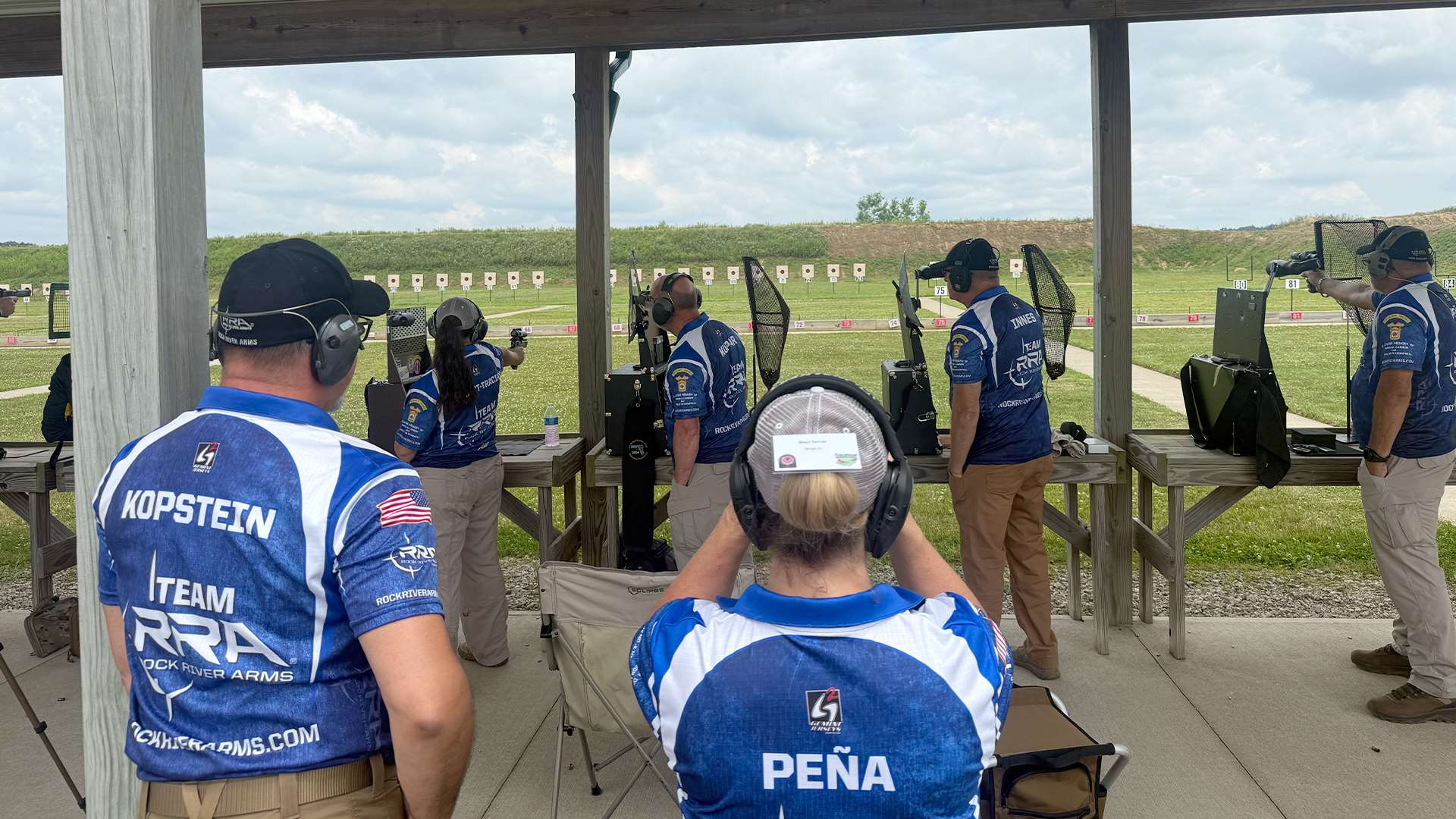 Team Rock River Arms shooters in blue and white jerseys compete in a centerfire team match at the Cardinal Shooting Center while teammates observe from behind the firing line