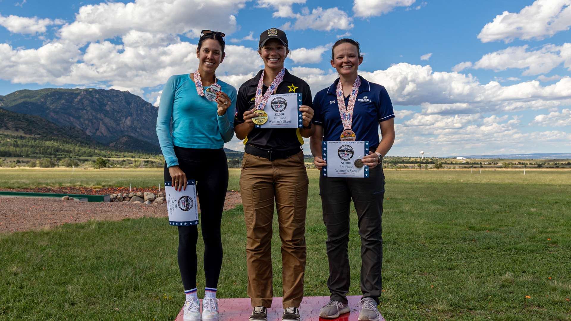 Three female skeet shooters stand on a podium holding medals and award certificates at the Fort Carson shotgun range