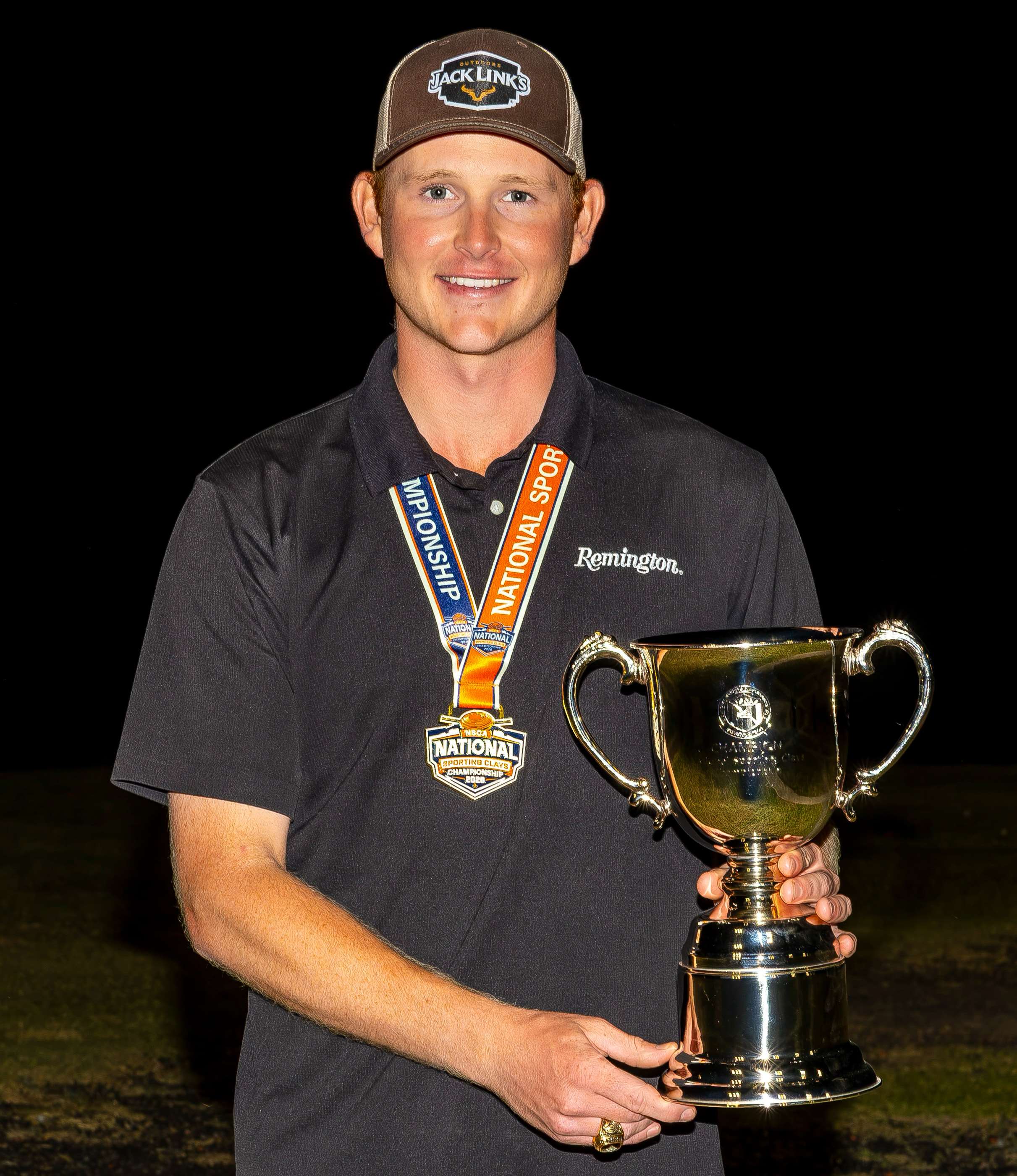 Smiling sporting clays shooter Todd Hitch poses at night with a two-handled silver championship cup and an orange-and-blue NSCA Nationals medal ribbon