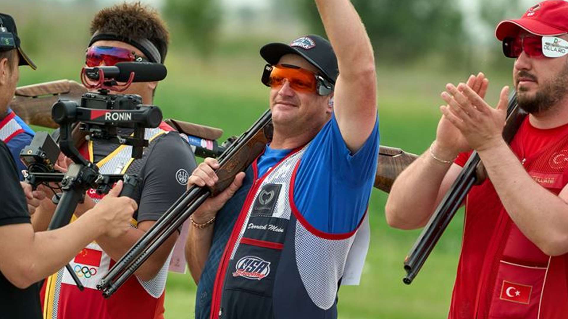 Derrick Mein raises his arm to acknowledge the crowd while standing between fellow Men's Trap finalists with his shotgun resting on his shoulder at the 2026 ISSF World Cup Shotgun in Almaty, Kazakhstan