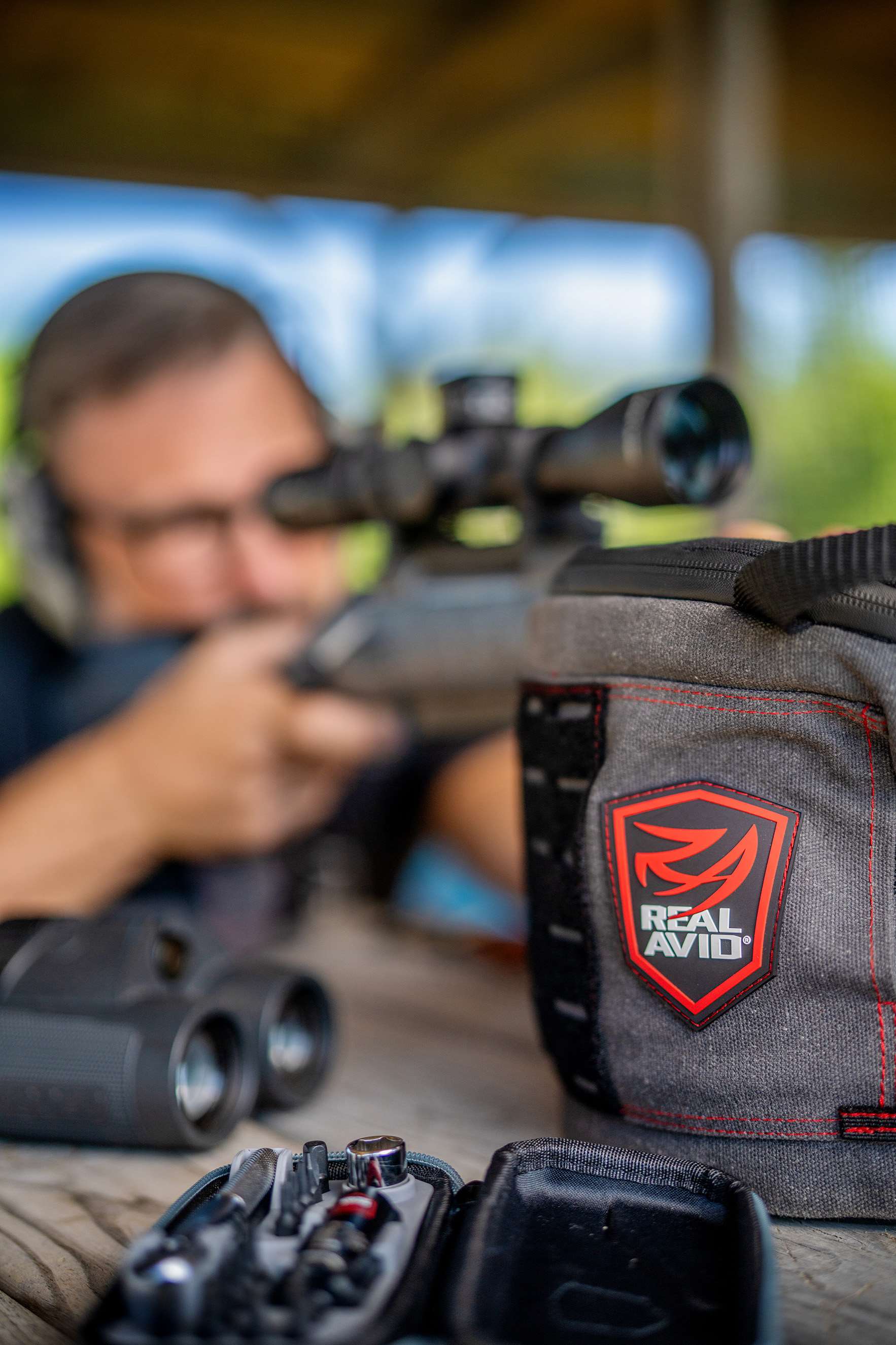 A scoped bolt-action rifle supported on a Real Avid shooting bag, with an open tool kit and binoculars in the foreground and pro shooter Doug Koenig behind the rifle