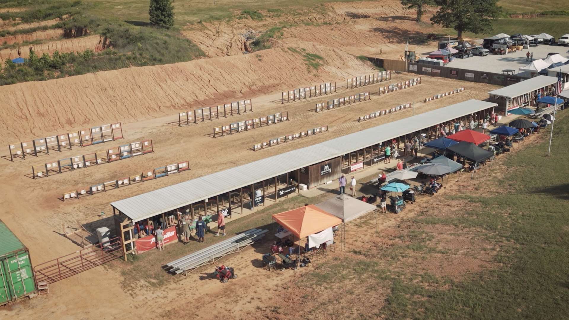 Aerial view of an outdoor shooting range: a long, covered firing line faces several rows of steel target lanes with multicolored frames set against a tall dirt berm. Spectators gather under pop-up canopies beside the line, and vendor tents and trucks line a gravel lot in the background