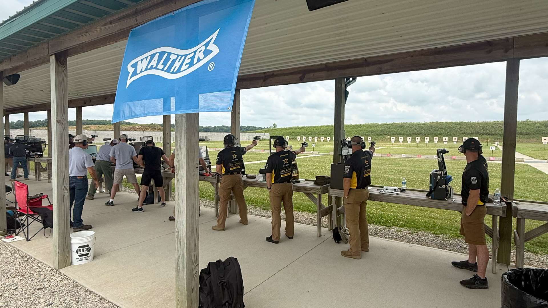 U.S. Army Marksmanship Unit shooters in black and gold team shirts fire pistols one-handed from a covered firing line beneath a blue Walther banner at the Cardinal Shooting Center