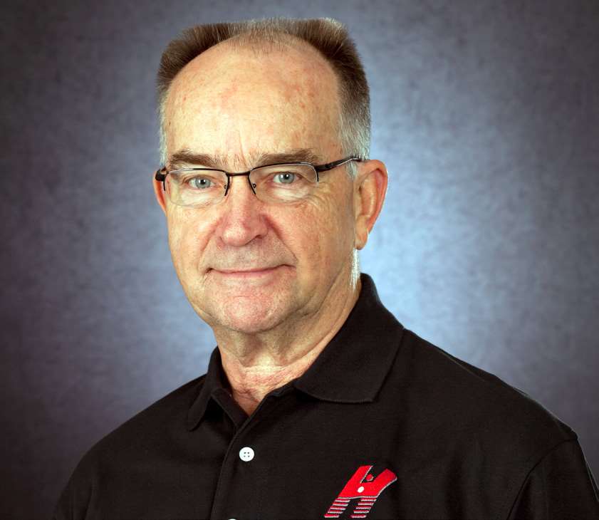 Studio portrait of gunsmith Richard Heinie wearing glasses and a black polo shirt with a red Heinie logo