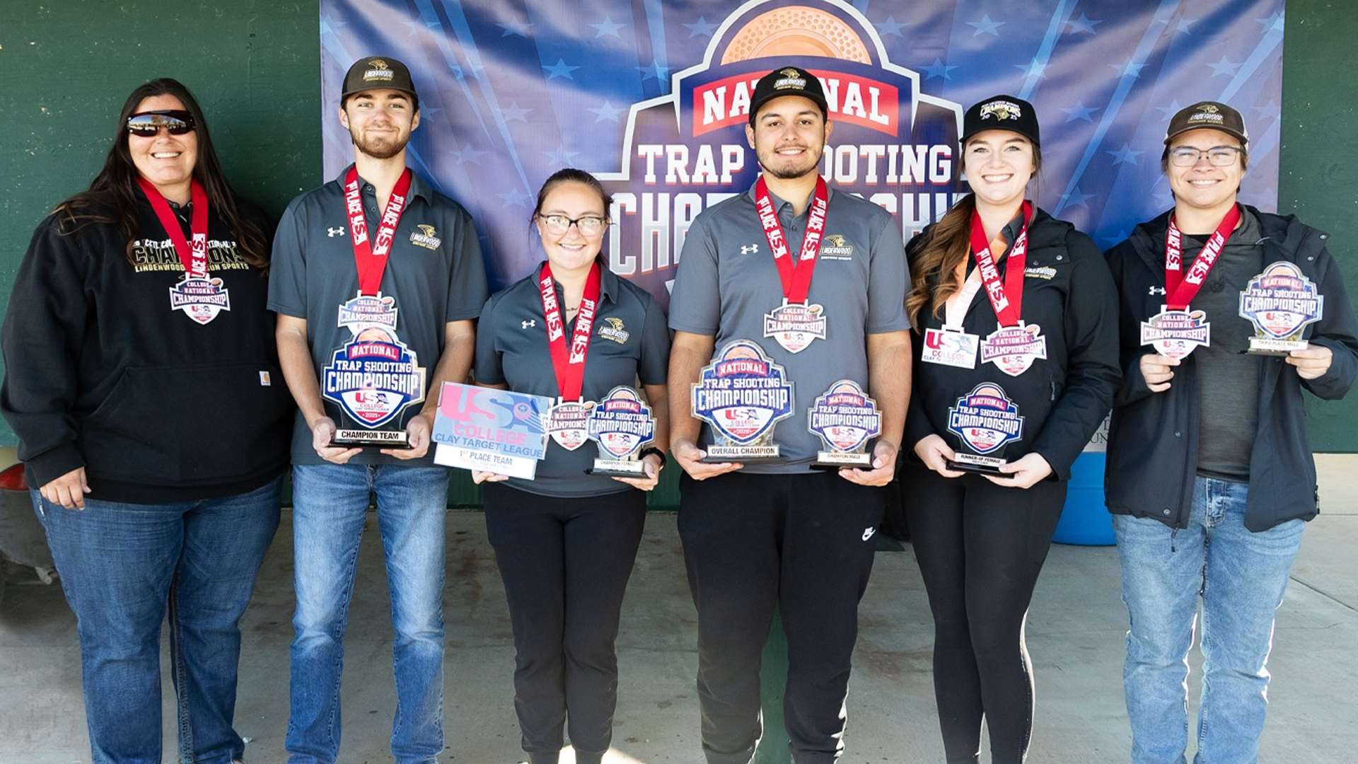 Six Lindenwood University shooters and a coach stand together holding trophies and wearing medals in front of a National Trap Shooting Championship banner