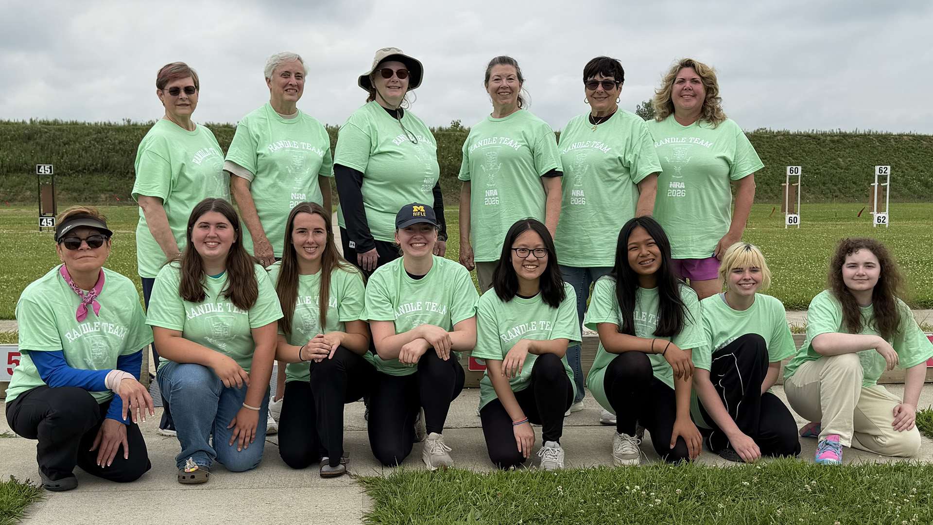 Fourteen women in light green Randle Team shirts pose in two rows on the firing line at the Cardinal Shooting Center