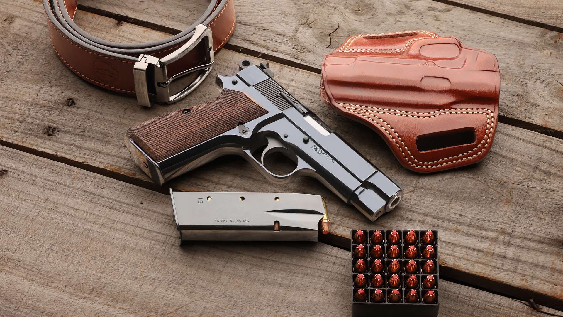 Right side of a polished blued Springfield Armory SA-35 4-inch 9 mm pistol on weathered wood planks beside a 15-round magazine, a box of hollow-point cartridges, a brown leather belt and a leather holster