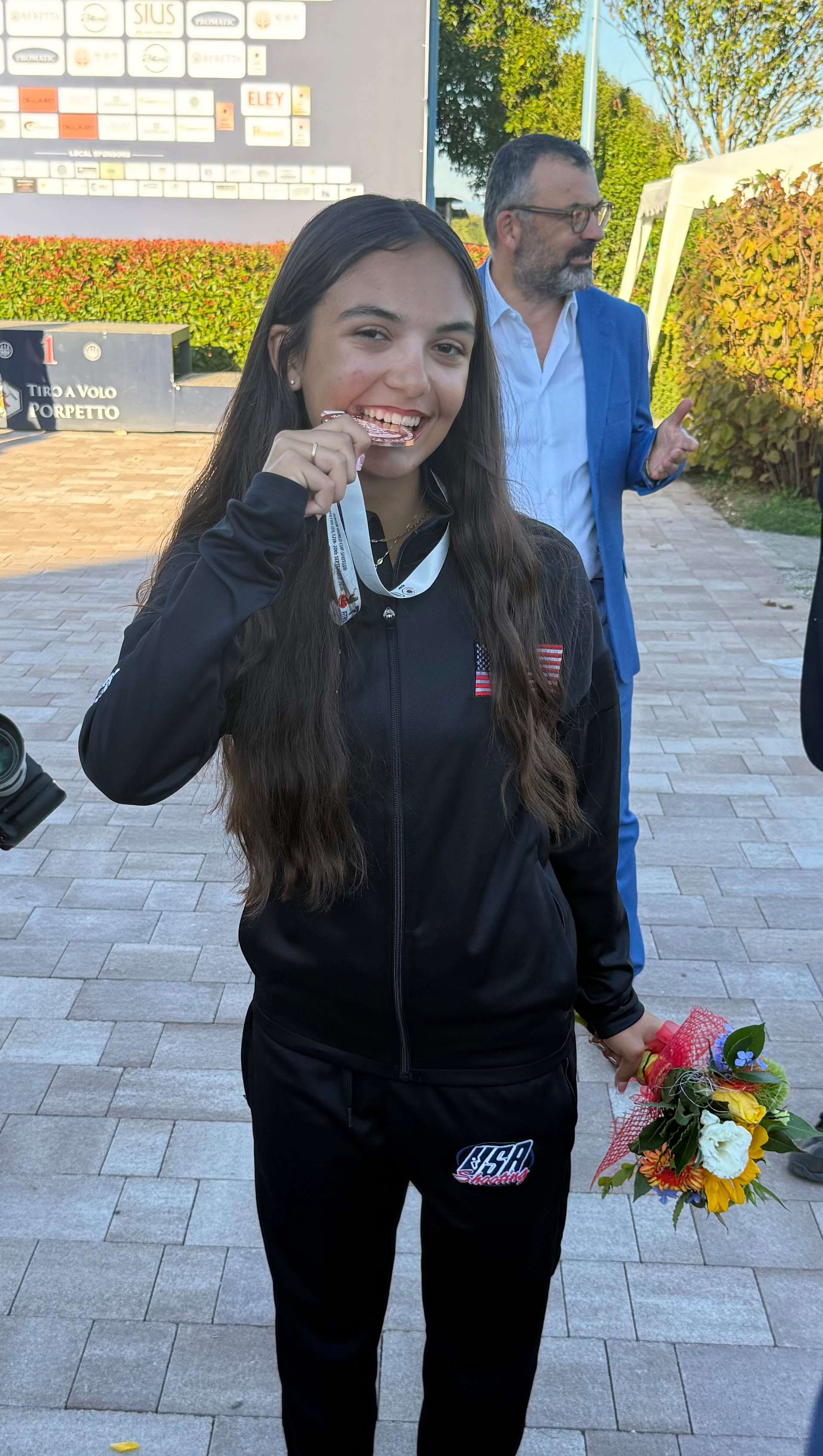 Amaris Cerra smiles and bites her bronze medal while holding a bouquet of flowers, with the Tiro a Volo Porpetto podium behind her