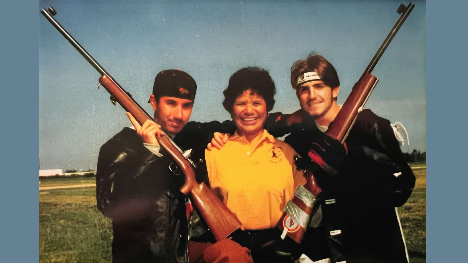 Joshua Keys and Aaron Tweedie stand in dark leather shooting coats holding smallbore rifles muzzle-up, flanking coach Leslie Cook in a yellow polo shirt