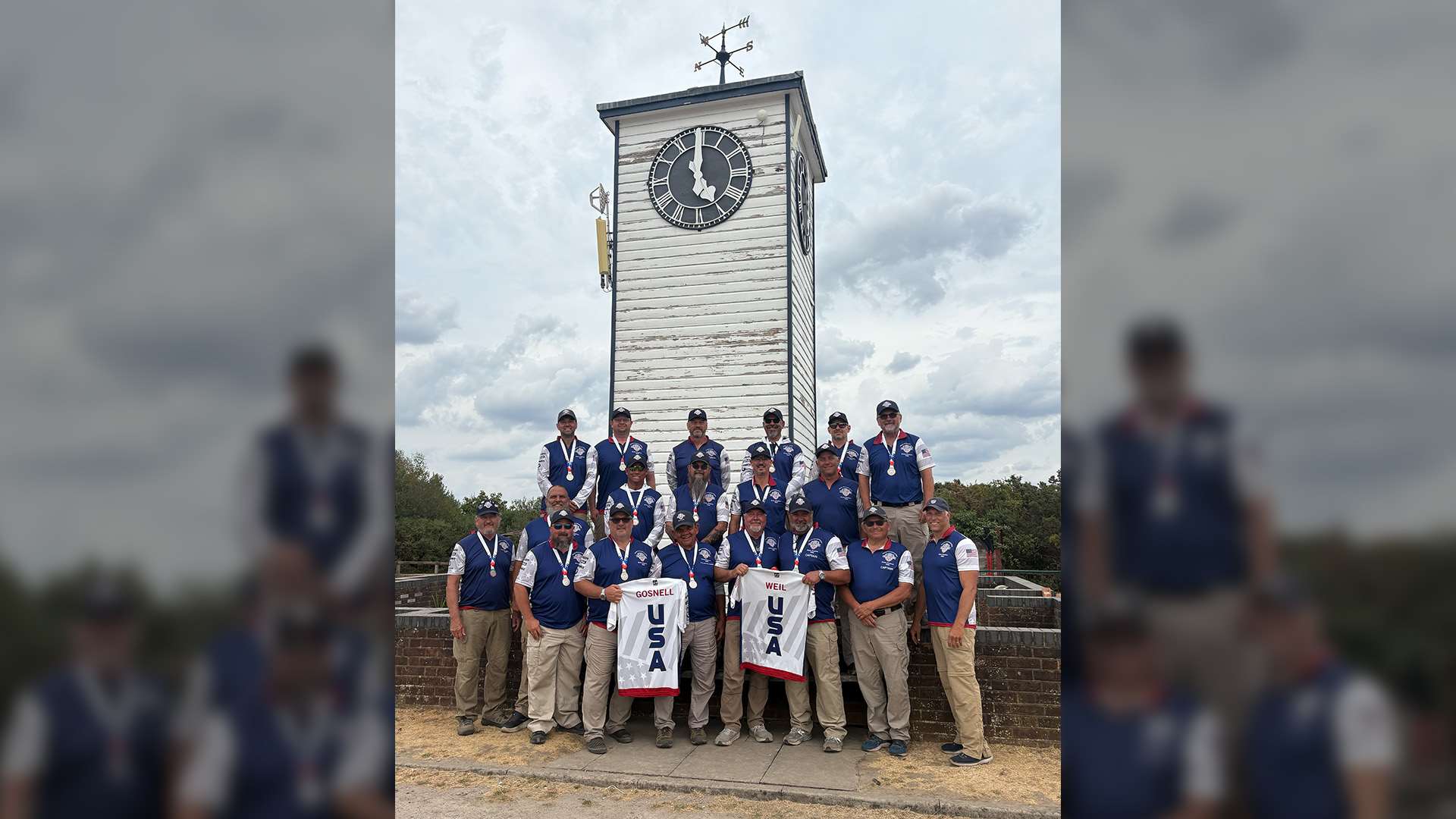 United States F-Class Open Rifle Team members wearing silver medals and team polos in front of the white clock tower at Bisley, two of them holding up USA jerseys bearing the names Gosnell and Weil