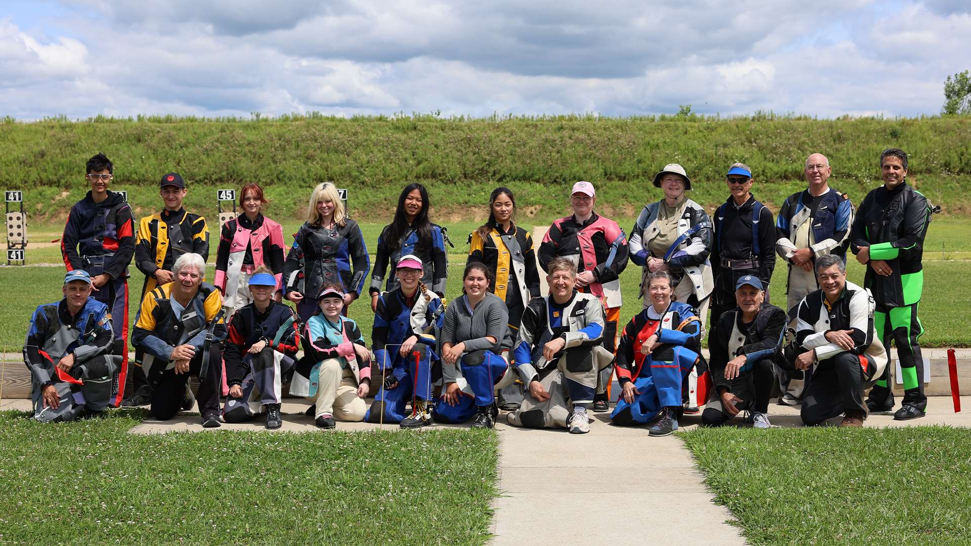 About two dozen smallbore rifle competitors in colorful shooting jackets pose in two rows on the firing line at the Cardinal Shooting Center, standing and kneeling in front of a grass berm lined with target frames