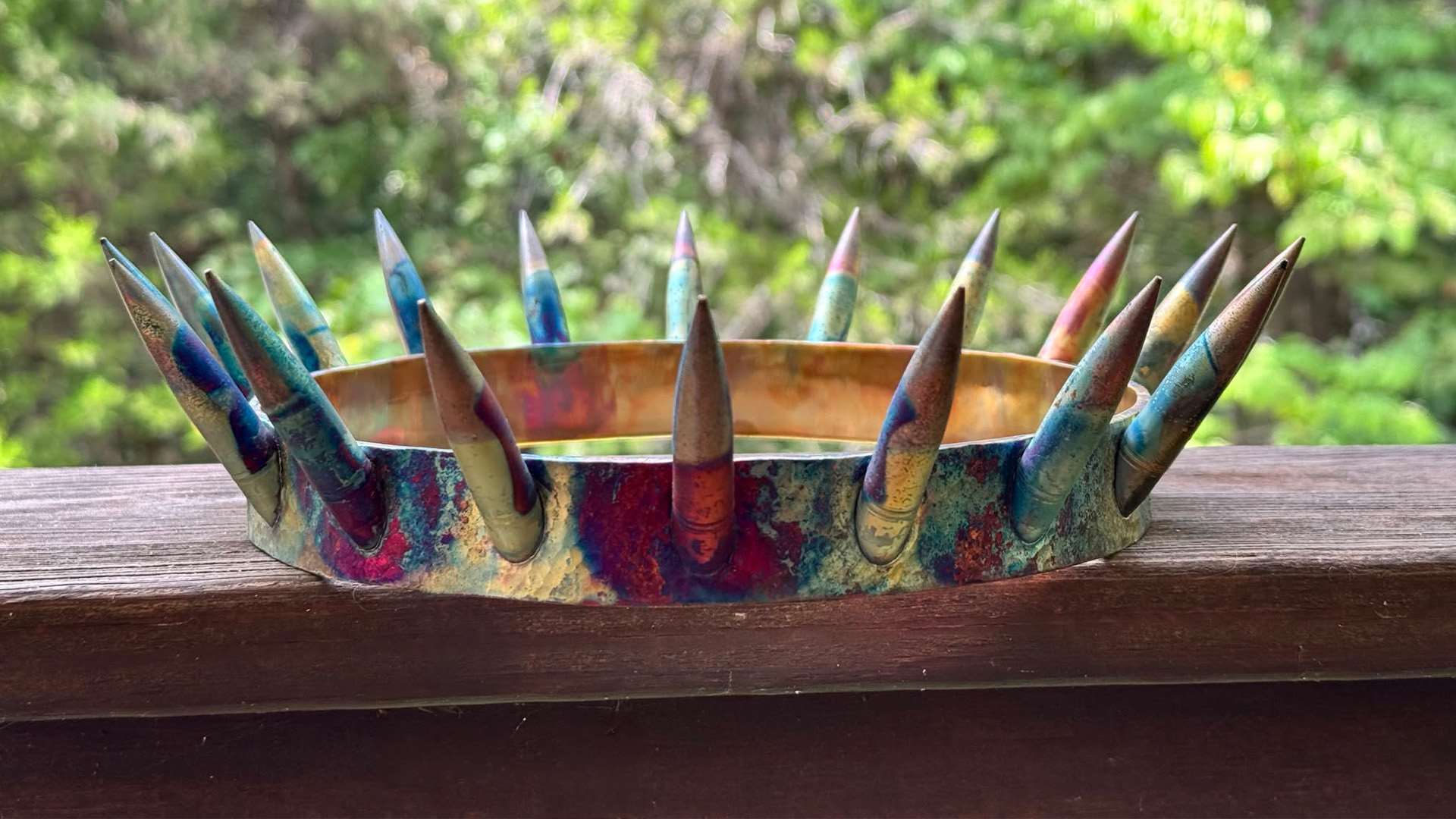 A crown with a multicolored metal band and a ring of upright bullet-shaped spikes, resting on a wooden railing with green foliage behind it.
