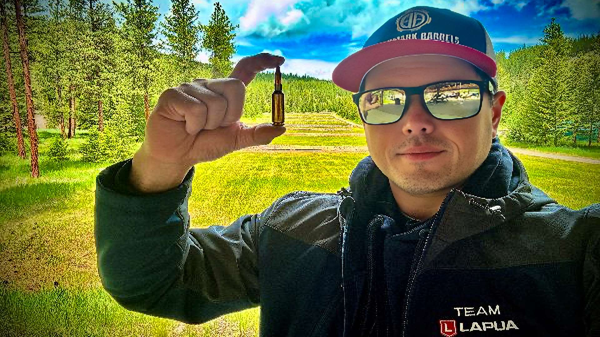 Erich Mietenkorte holding up a single centerfire rifle cartridge between his fingers at a shooting range, wearing sunglasses, a Bartlein Barrels cap and a Team Lapua jacket